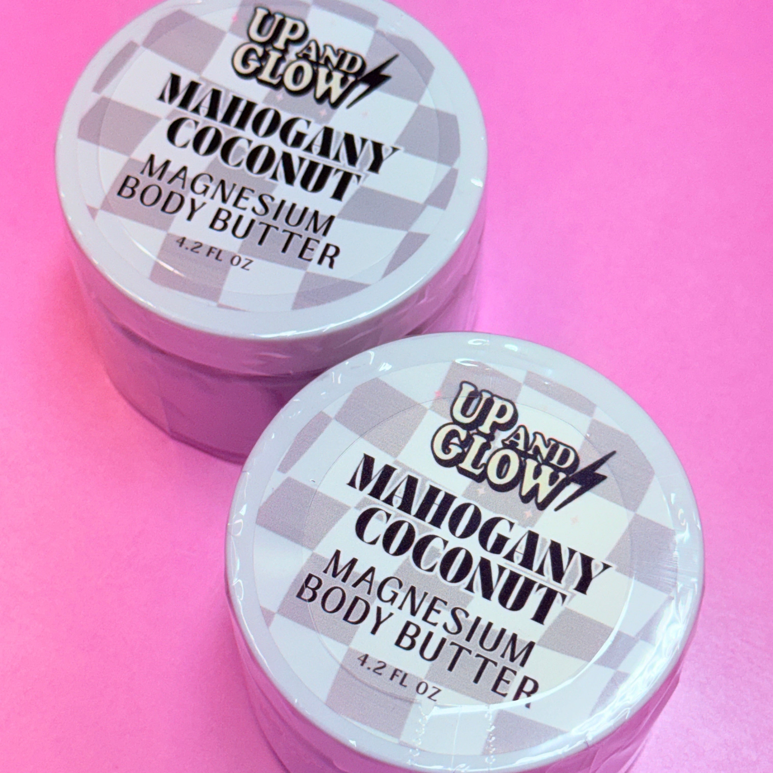 Two containers of Up & Glow Mahogany Coconut Magnesium Body Butter on a pink background